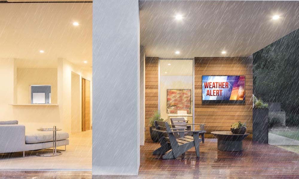 A modern outdoor living space during a rainstorm, featuring a weatherproof TV displaying a “Weather Alert” message, mounted on a wooden wall under a covered patio with seating.